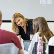 A lecturer smiling while stood at the front of a classroom.