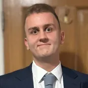 Daniel, a young man with brown hair and eyes and wearing a navy blue blazer and grey tie, smiles at the camera