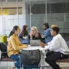 A group of three people sat around a table. They're talking in the group with a share laptop in the middle.