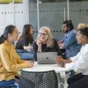 Three people sat a desk talking. The person in middle has a laptop open on the desk.