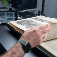 A close-up of a person's hands working with an open historical book on a specialized scanning table. One hand is turning a delicate page with illustrations, while the other holds a tablet displaying a digital scan of the same page. The workspace includes modern scanning equipment and cables in the background, blending traditional and digital technologies.