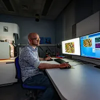 A technician works at computers displaying microscopic images, with a TESCAN electron microscope nearby.