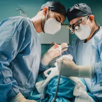 Two men in a surgery theatre. They're wearing scrubs, safety masks, and hats. They're holding a medical operating tool.  