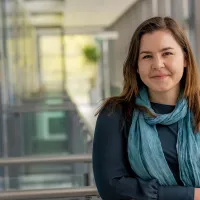 A young woman in a dark blouse and blue scarf smiles confidently in a modern university hallway.