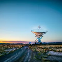 A towering satellite dish positioned in a wide, grassy field, silhouetted against a vivid orange sunset sky, with a winding dirt road leading toward it.
