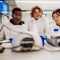 Three students in lab coats and safety goggles working together in a science laboratory.