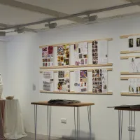 An art gallery room of the MA Contemporary Curating final show of 2024. There are hanging sculptures and design boards on the wall, plus tables displaying printed work.
