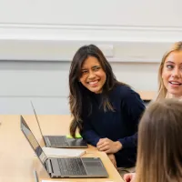 A group of students smiling having a discussion. There are open laptops on the table in front of them.