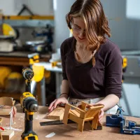 Student wearing safety glasses works on a wooden model at a workshop bench.