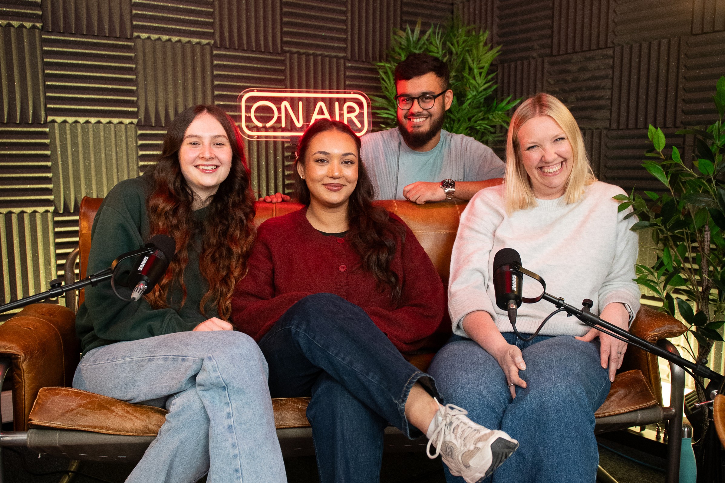 A group of four people sat on a sofa in a podcast recording studio, in front of microphones with a neon 'on air' sign behind them.